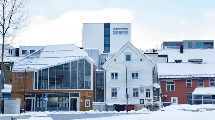 Building with snow-covered roof stands in a snowy environment. Signs read "Comfort Hotel Xpress," "Rønning Hansen," and "CENTIMETER" on the wooden facade.