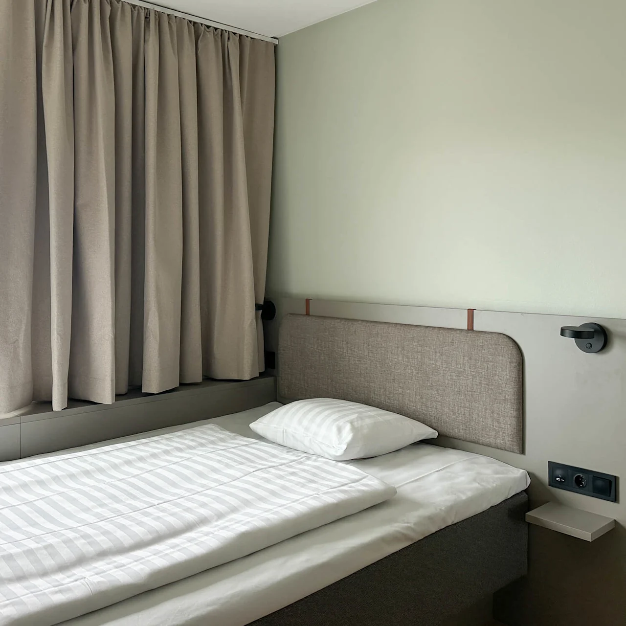 A neatly made single bed with striped bedding is beside a large window with beige curtains, situated in a minimalist room with light-colored walls.