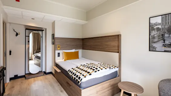 Et koselig hotellrom med en enkeltseng, sengegavl i tre og et speil nær inngangen.