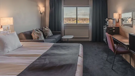 A neatly arranged hotel room features a bed with white linens, a gray sofa with decorative pillows, a desk with a chair, and a large window overlooking a scenic landscape.