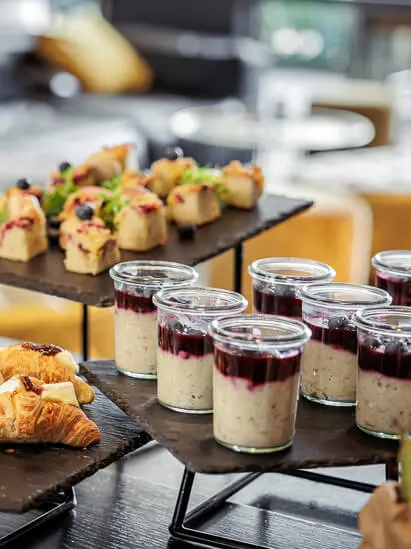 Et utvalg av appetittvekkere, inkludert croissanter, smørbrød, fargerike desserter i glasskrukker og fruktspyd, er elegant utstilt på en svart skifer i en lys, uformell spiseinnstilling.