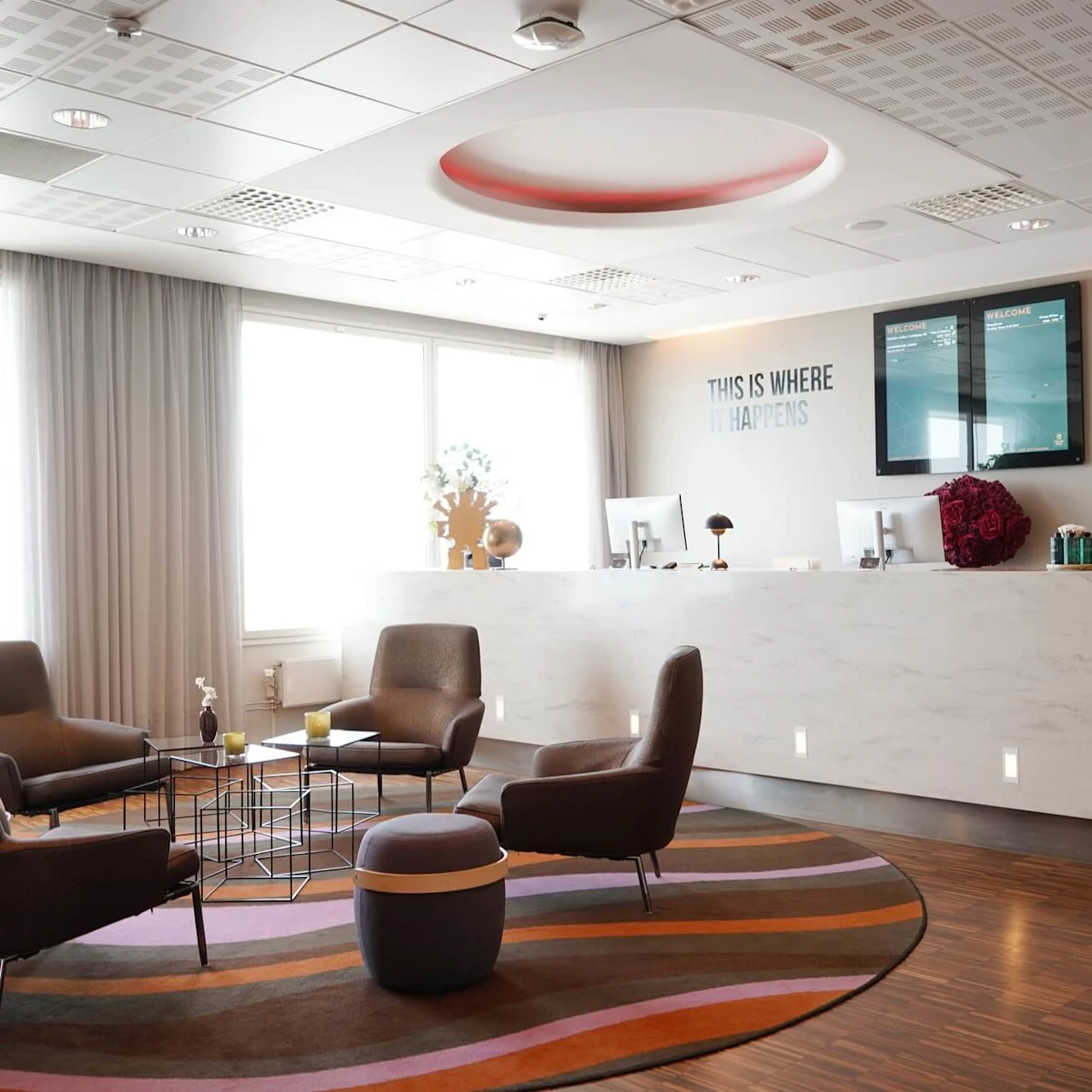 Reception area with four chairs around tables on a colorful rug. White desk with computers behind, wall reads "THIS IS WHERE IT HAPPENS." Modern decor and lighting.