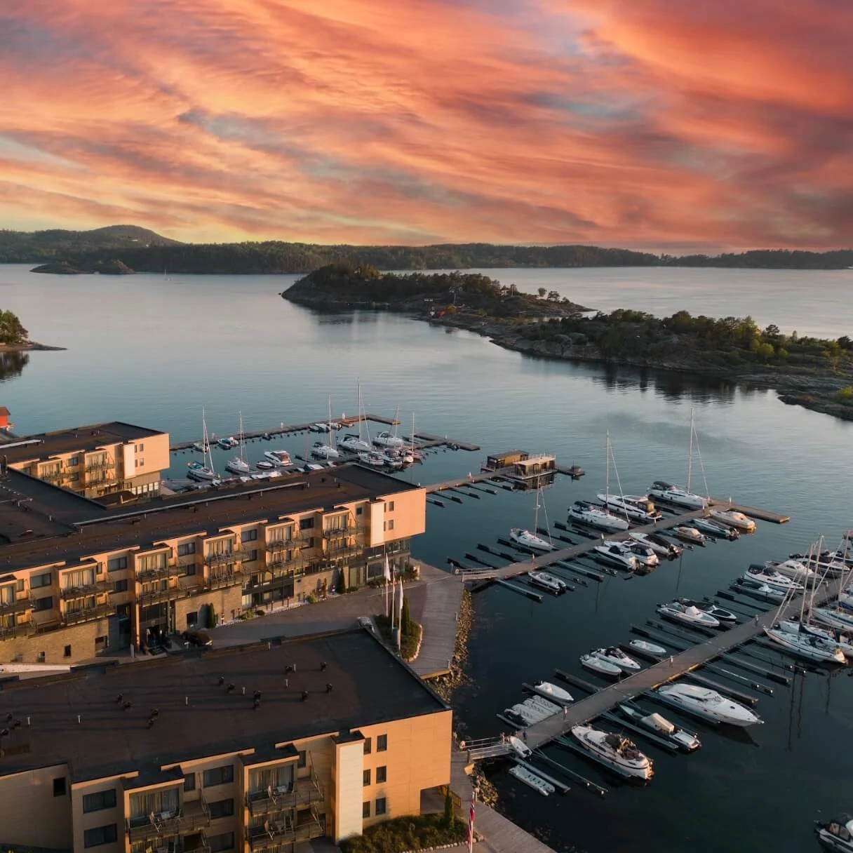 Moderne bygninger har utsikt over en marina fylt med fortøyde båter mot en bakgrunn av åser og en rolig innsjø. Himmelen er malt med livlige oransje og rosa skyer.