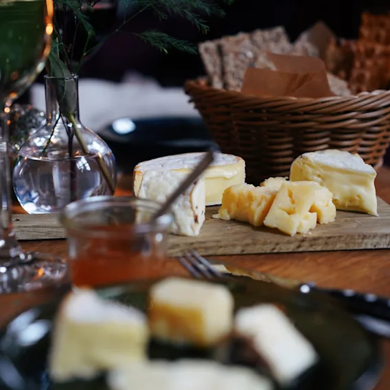 Osteutvalg hviler på et trebord med brødkurv og vin i nærheten; satt på et middagsbord med en vase med løvverk i et svakt opplyst miljø.