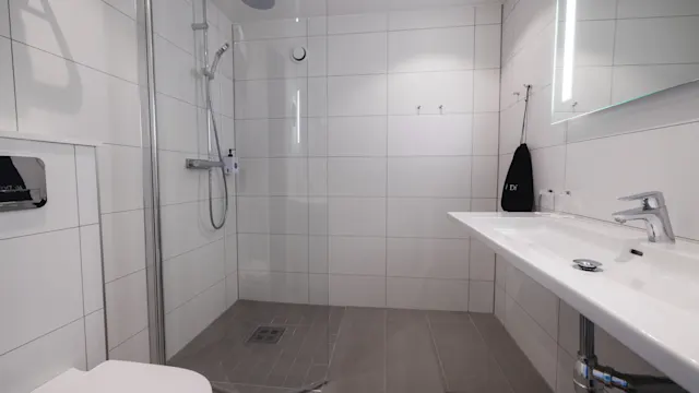 Ett modernt badrum med glasdusch, väggmonterad toalett och lång handfat i en ren, vitt kaklad miljö med grått klinkergolv. En handduk och toalettartiklar kompletterar den minimalistiska designen.