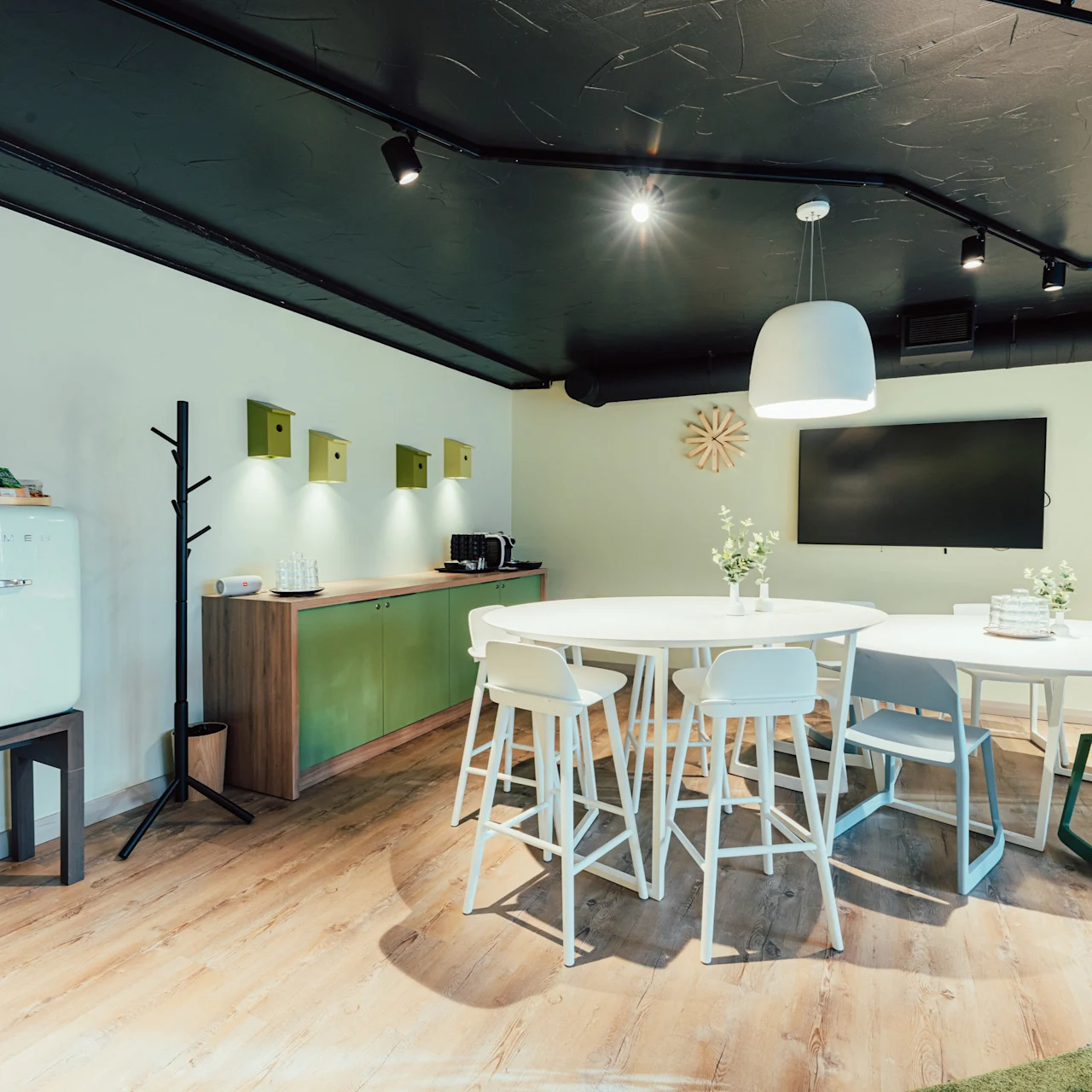 A round table surrounded by white chairs stands on wooden flooring under a white ceiling light. A black monitor hangs on the wall, and a retro fridge is placed nearby.