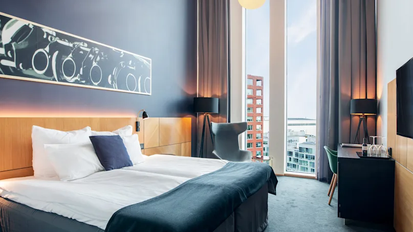 A bed with white linens is positioned in a modern hotel room with a large window; decorative art and furniture create a stylish atmosphere.