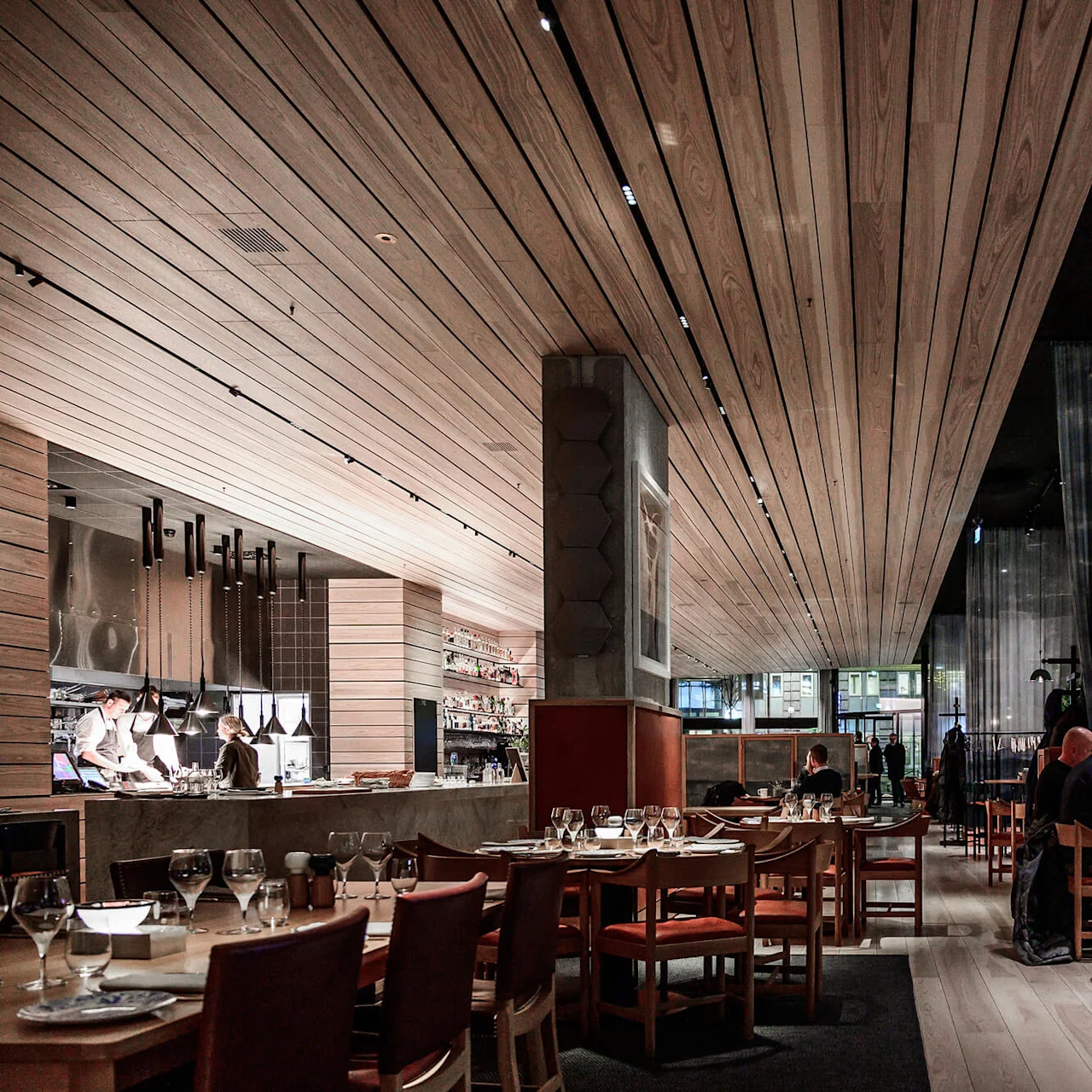 A modern restaurant with wooden interiors features people dining at tables with wine glasses. Waitstaff prepare meals at an open kitchen, contributing to an elegant, lively atmosphere.