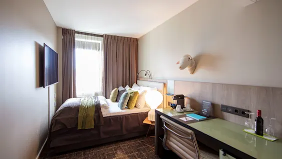 A cozy hotel room features a neatly made bed with green and white pillows. A desk holds a lamp and coffee maker. A window with curtains provides natural light.