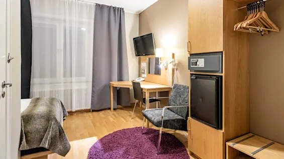 Et ryddig hotellrom med et skrivebord og stol i tre, foran en veggmontert TV. Et nattbord med lampe ved siden av en liten garderobe med kleshengere. Lilla teppe på tregulv.