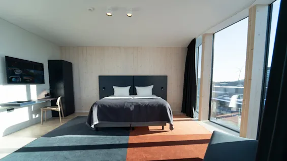A modern hotel room features a neatly made bed with white pillows, situated at its center. A large window offers a cityscape view, while a TV and desk occupy one side.