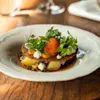 Gourmet-annos tuoreilla yrteillä, oranssinvärisellä ruoalla ja perunoilla, tyylikkäästi tarjoiltuna valkoisella lautasella puupöydällä.