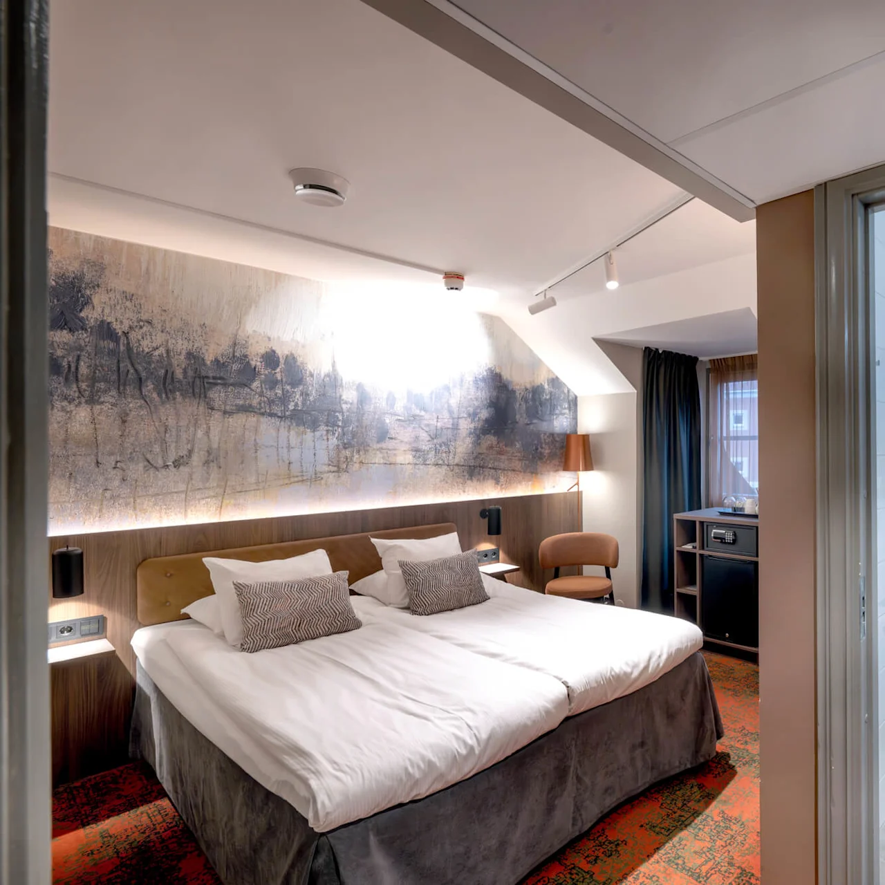 A hotel room with two beds and abstract wall art, adjacent to a bathroom featuring tiled walls and a hanging toilet; a small window provides ambient light.