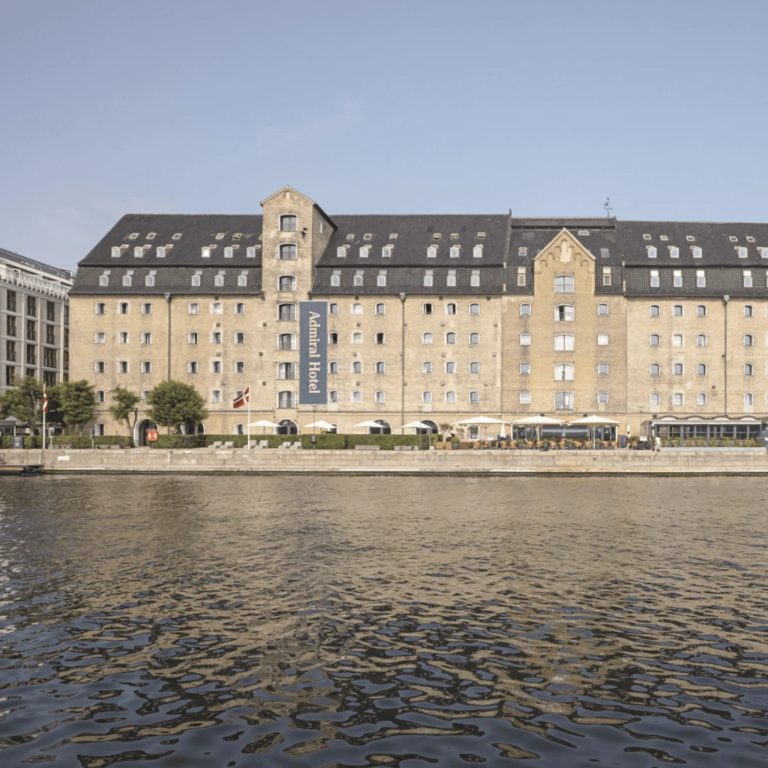 A large historic brick building faces a calm waterfront under a clear sky. An attached banner reads: "Admiral Hotel." Trees and a boat are visible nearby.