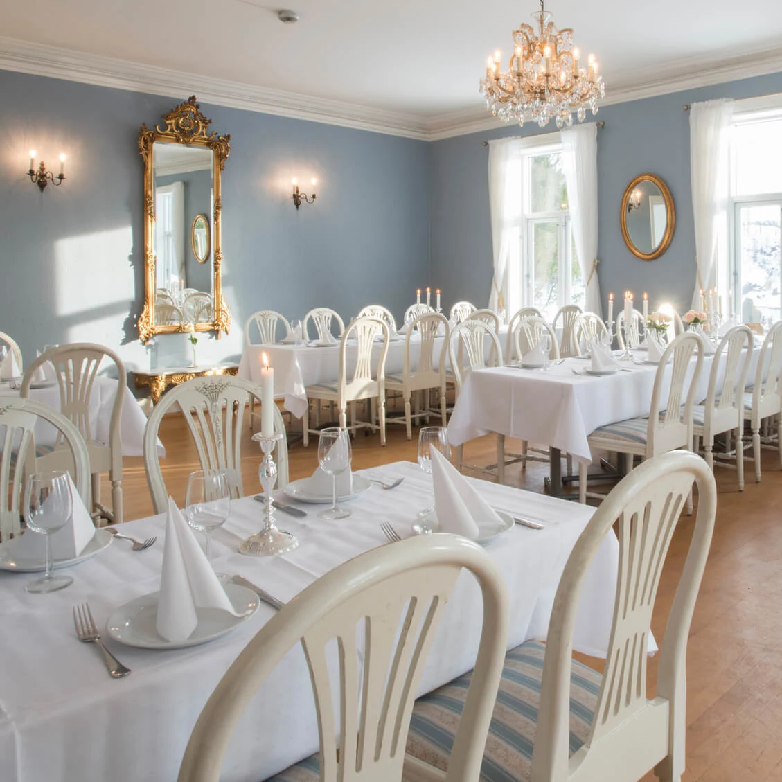 Elegant matsal med dukade bord, kristallkronor, speglar, på en solig dag med utsikt över snötäckt terräng utanför.