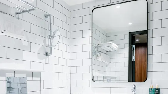 Et lyst baderom med fliser på veggene. Et rektangulært speil over en vask reflekterer hyller med håndklær. Et toalett er ved siden av. Ingen tekst er til stede.