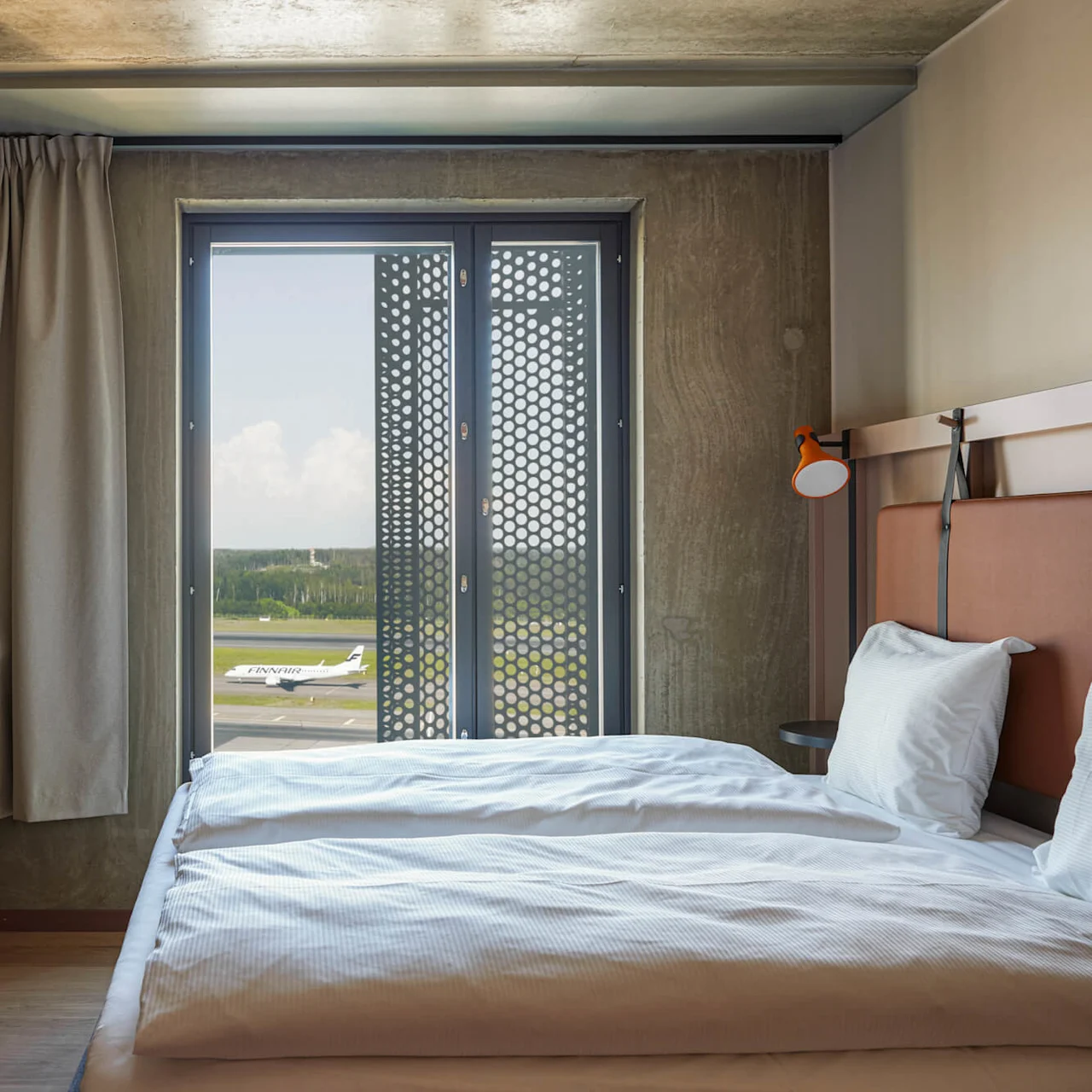 Et moderne hotelværelse med to enkeltsenge og udsigt over en landingsbane gennem et stort vindue. Gardinet dækker delvist vinduet og afslører et fly med "Finnair."