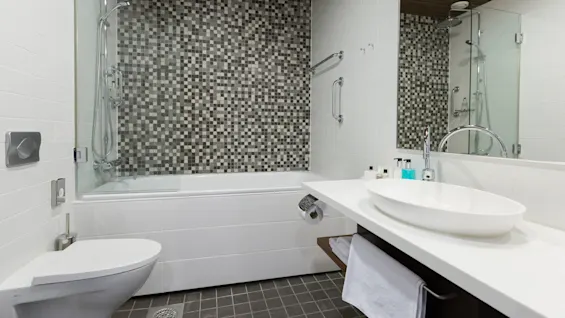 A modern bathroom with a mosaic-tiled wall behind the bathtub, a large mirror, and a sleek sink counter.