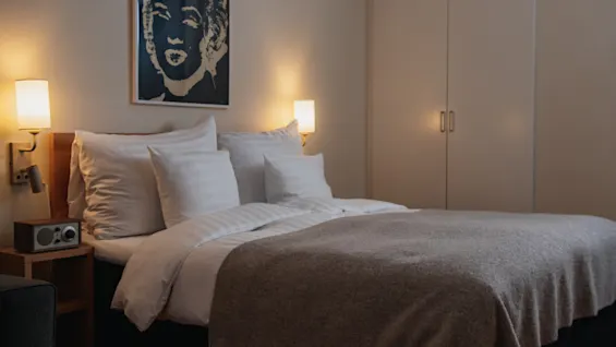 A neatly made bed with multiple pillows is illuminated by two wall-mounted lamps; a dark abstract portrait hangs above the bed in a cozy room with a wardrobe.
