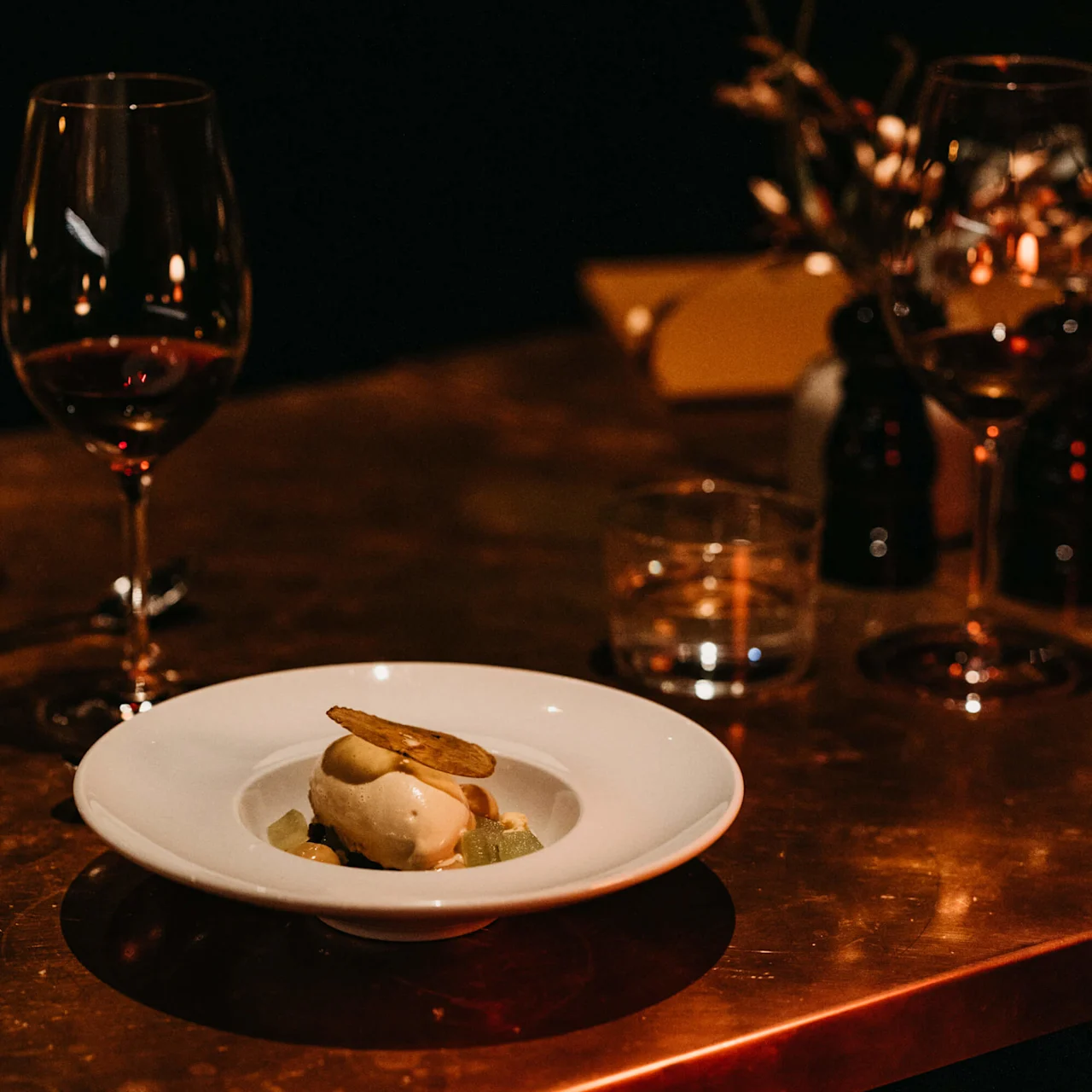 A gourmet dish on a plate with an elegant dining backdrop, including wine glasses and soft lighting.