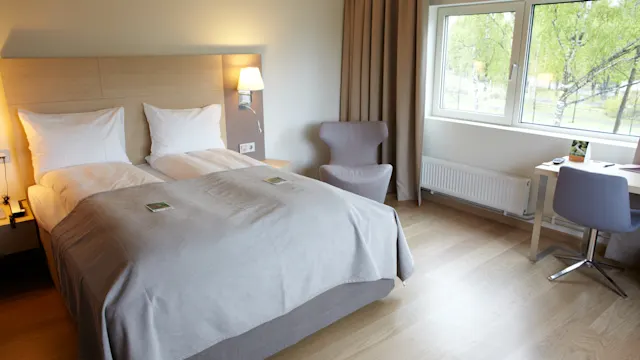 En pent oppredd seng med hvite puter og et grått teppe står ved siden av en tent vegglampe. En stol og et skrivebord står nær et vindu med trær synlige utenfor.
