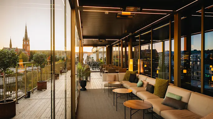 En moderne terrasse med koselige sitteplasser har nøytrale sofaer og små bord. Utsikt over et bylandskap med en fremtredende kirke og trær, innrammet av store glassvinduer.