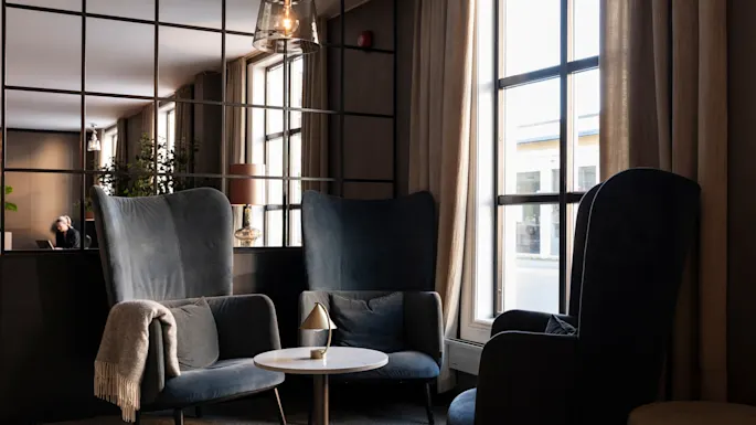 A comfortable lounge space featuring plush armchairs, a small table, and a bright window, with a person working nearby.