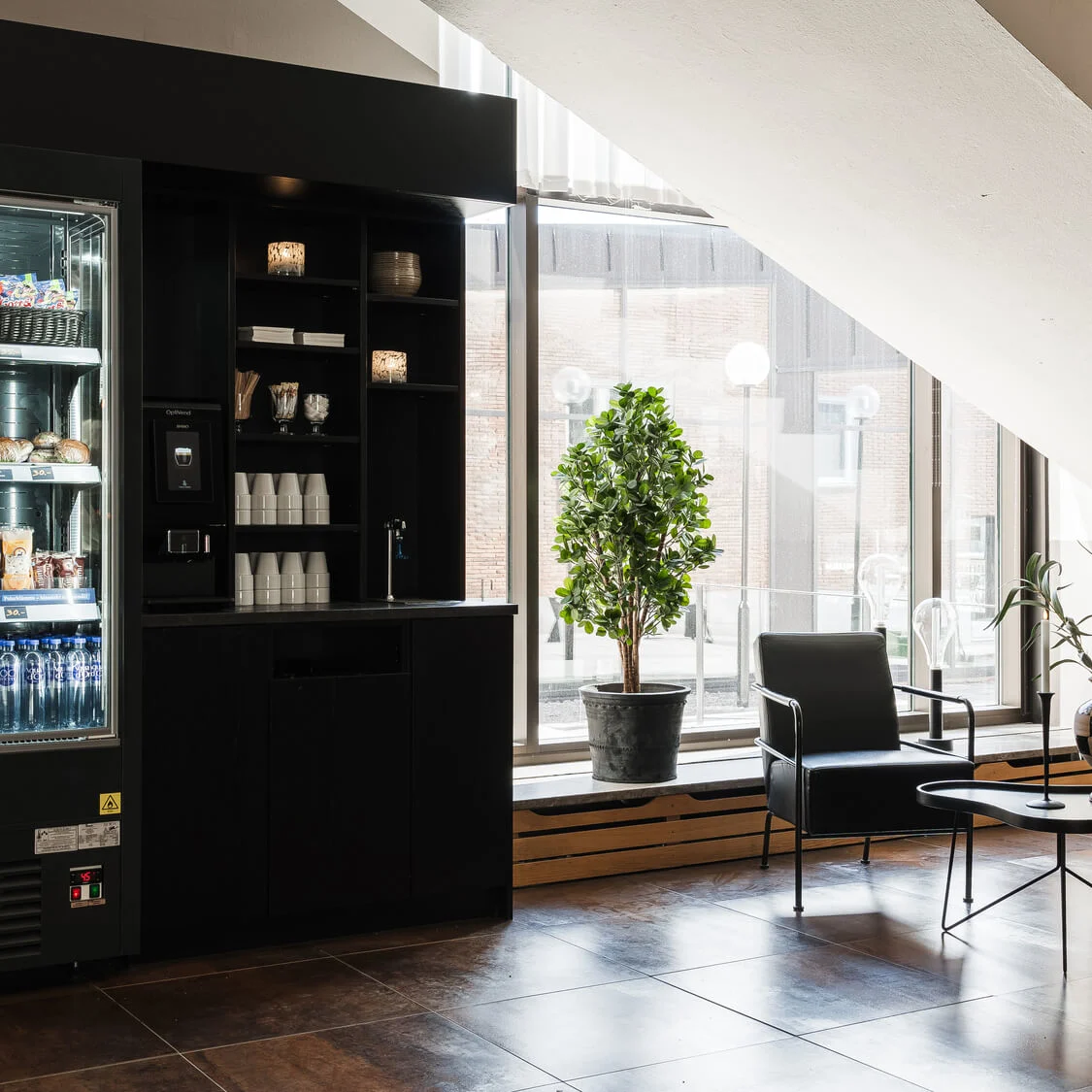 Varuautomat visar snacks och drycker. Nära, stolar och ett bord med en växt skapar en sittgrupp under en trappa. Stort fönster släpper in naturligt ljus.