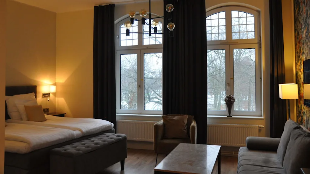 Et velutstyrt rom med en seng, sofa, og lenestol, store vinduer med mørke gardiner og utsikt til trær.