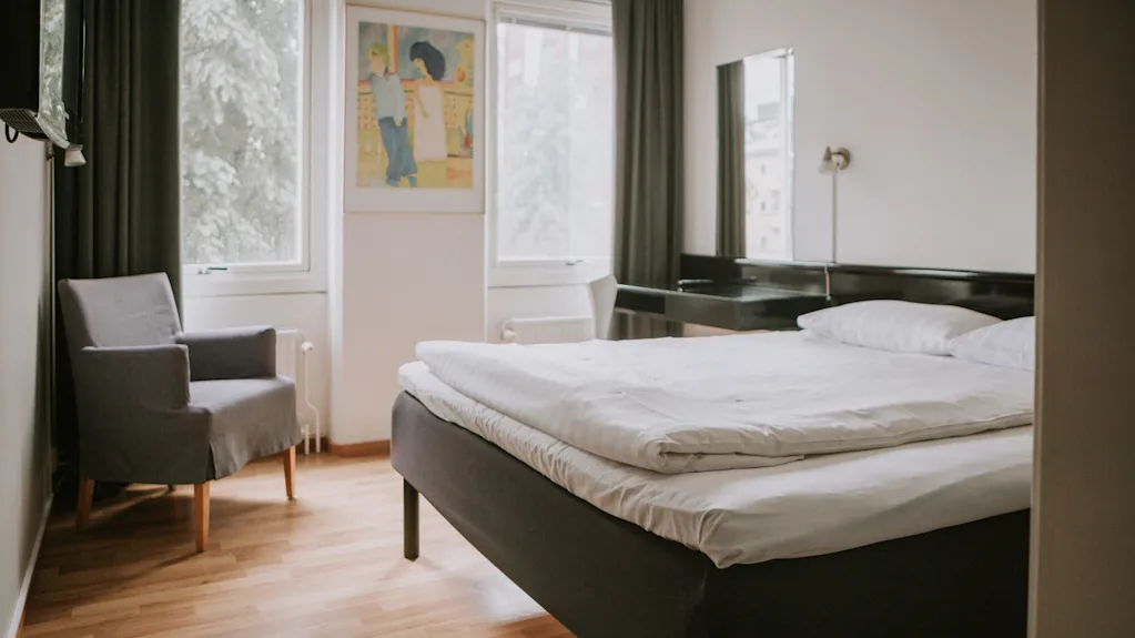 En pænt redt seng med hvidt sengetøj står ved siden af en grå lænestol i et rum med trægulv, et vægmaleri, store vinduer og en kommode med spejl.
