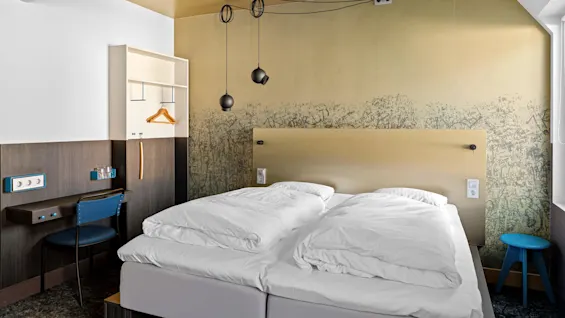 En pent oppredd dobbeltseng står i et moderne, minimalistisk rom, med en mønstret gul vegg, hengende svarte pendellamper, et lite skrivebord med stol og en hylle med hengere.