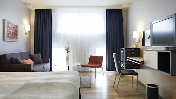 A modern hotel room has a bed, sofa, table, and chair with natural light from large windows. Dark curtains frame the room, complemented by a wall-mounted TV and minimal decor.