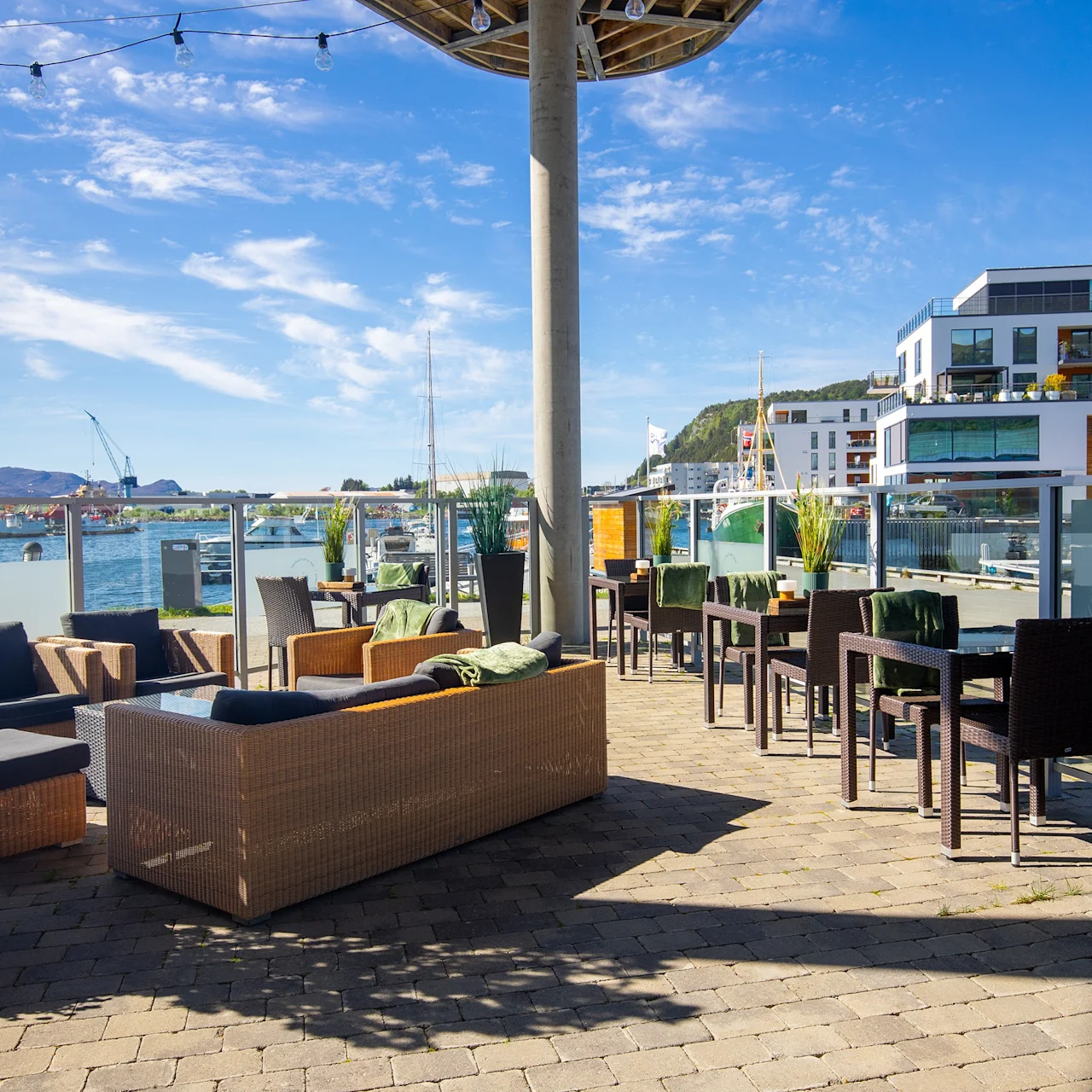 Utendørs terrasse med rottingmøbler, bord og stoler under lysslynger. Utsikt over en havn med moderne bygninger i bakgrunnen, omgitt av klar blå himmel og grønt.