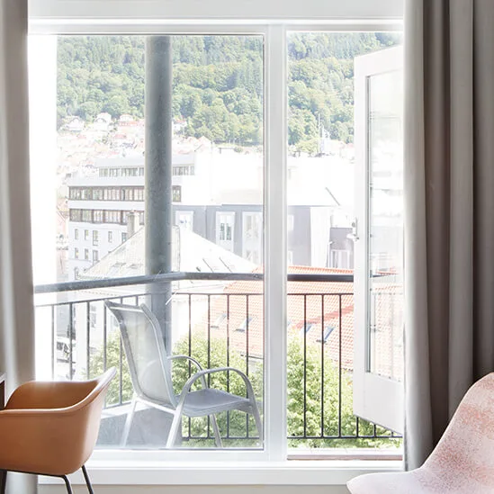 Et lyst hotelværelse med skrivebord, lampe, stole og et vægmonteret TV. En åben dør fører til en balkon med udsigt over hustage og grønne bakker.