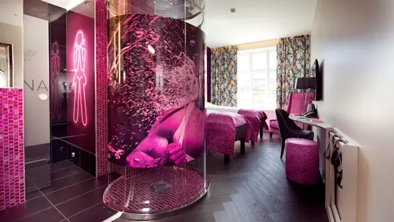 A circular shower with vibrant pink decor features a large design and is placed near twin beds covered with purple duvets in a modern hotel room with floral curtains.