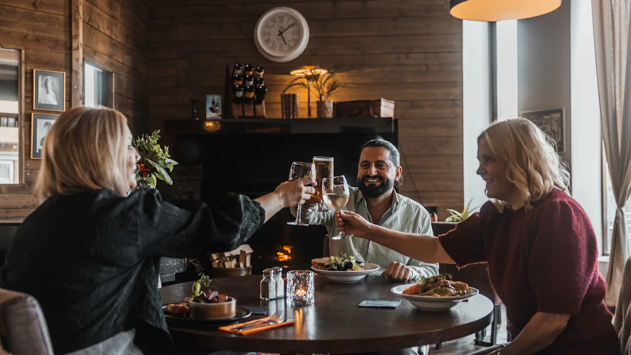 Kolme ihmistä kohottaa maljoja juomien ja ruoan kera viihtyisässä ravintolassa tai baarissa, nauttien sosiaalisesta tapaamisesta.