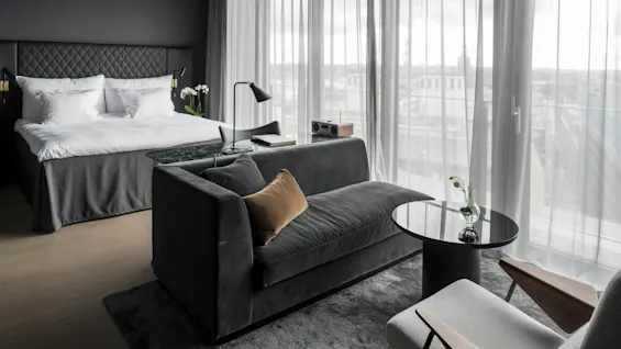 Et ryddig hotellrom har en stor seng, en mørk sofa og et glassbord med blomster, innrammet av gulv-til-tak-vinduer med tynne gardiner.
