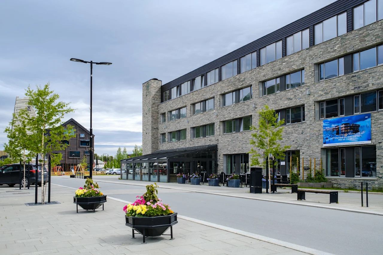 En moderne steinbygning langs en rolig gate, pyntet med fargerike blomsterkasser, flankert av unge trær og et stort banner. Overskyet himmel og gatelykter gir urban stemning.