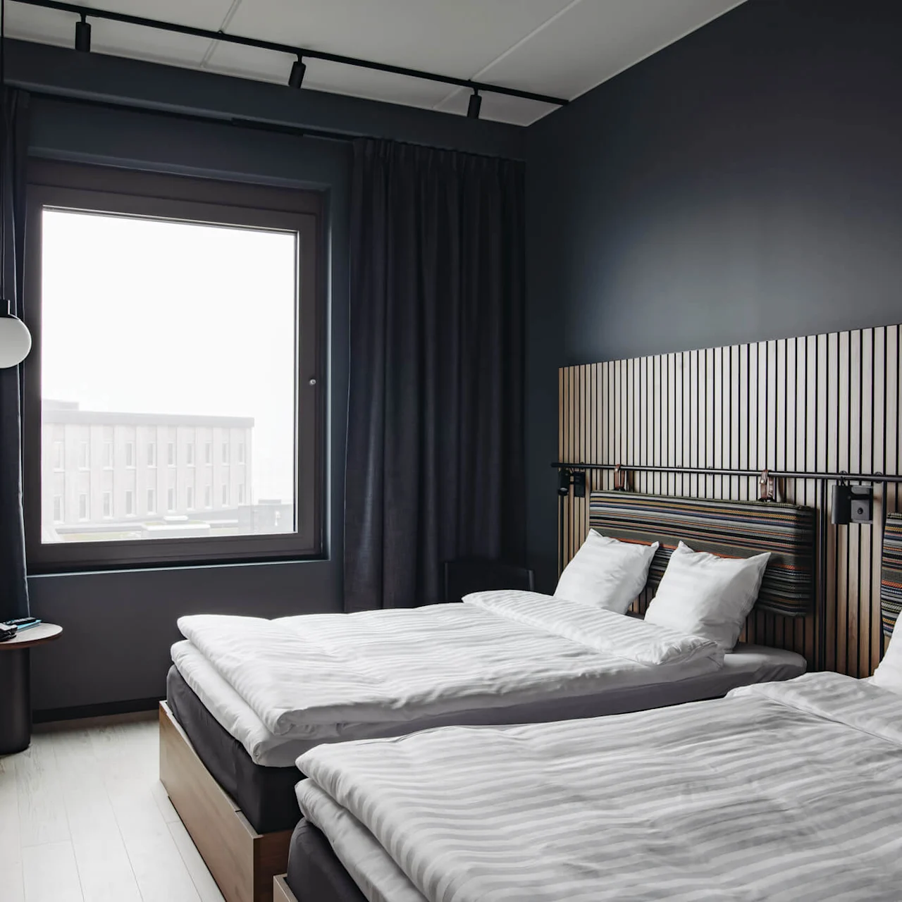 Two neatly made beds, topped with white linens, rest in a modern room. A large window reveals an urban building view, surrounded by dark walls and curtains.
