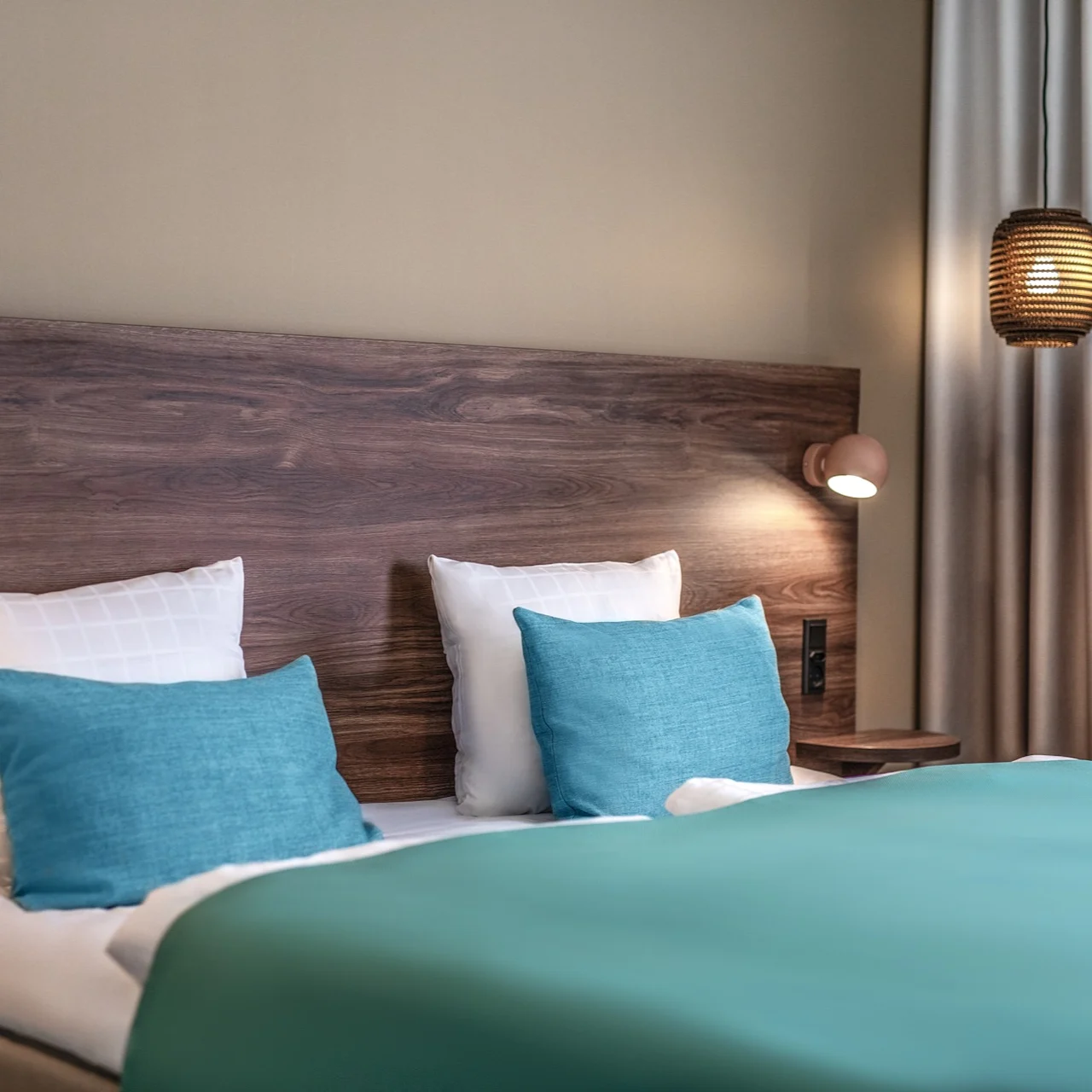 A bed with teal pillows and blankets sits against a wooden headboard. A small wall lamp and hanging light illuminate the bedroom, with beige curtains on the right.
