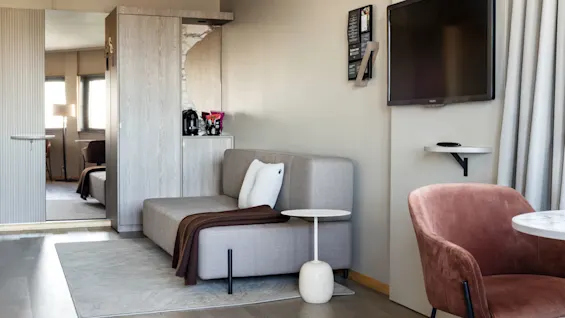 A grey sofa with pillows and a blanket sits beside a small white table. Nearby, a wall-mounted TV and a velvet chair complete the elegant, modern room.