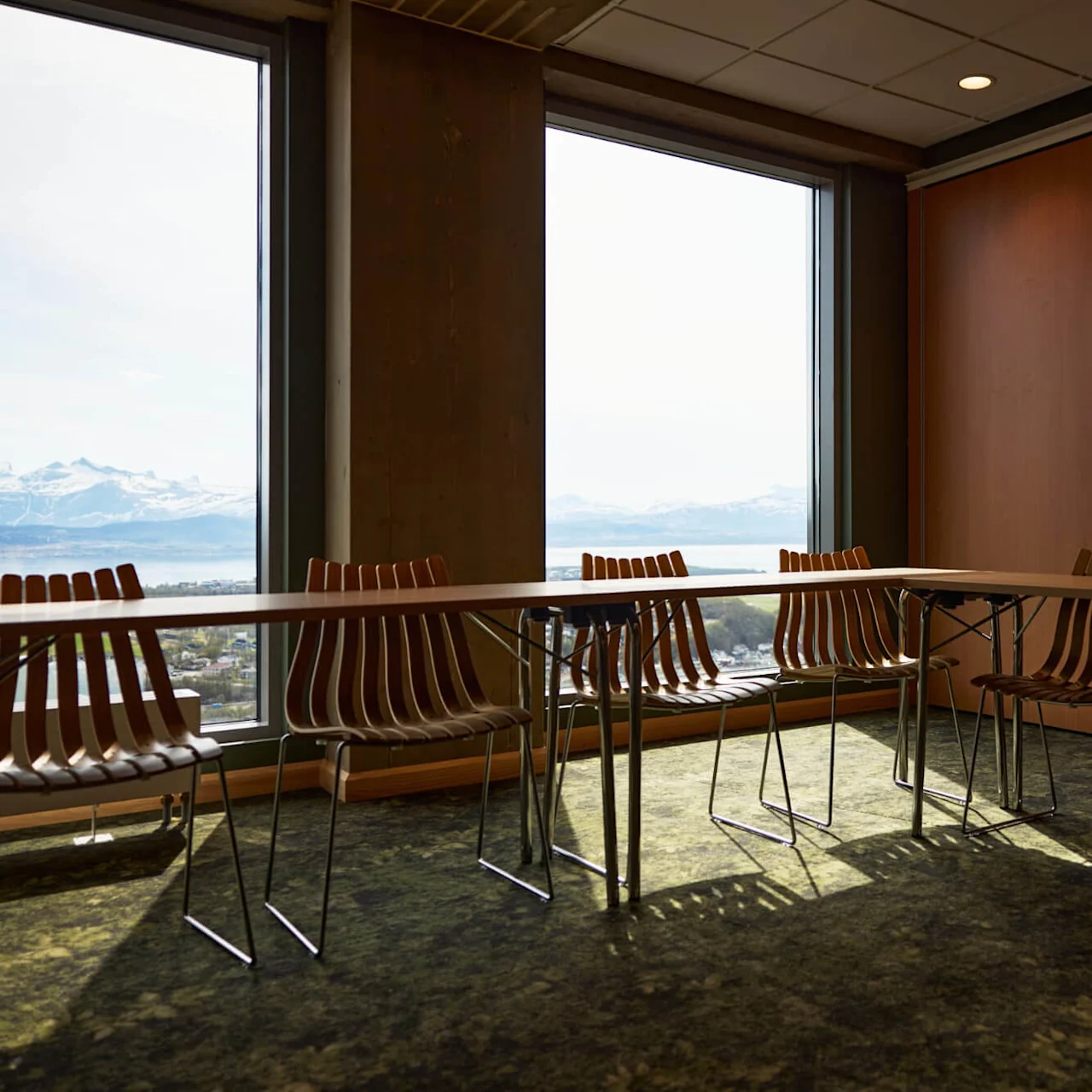 Tables and chairs are arranged in a well-lit room with large windows offering a view of snow-capped mountains. The walls are wood-paneled, and the floor has a patterned carpet.