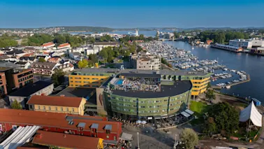 En grön och gul byggnad med takfester blickar ut över en marina full av båtar, omgiven av en kuststad med färgglada hus och frodig grönska under en klarblå himmel.