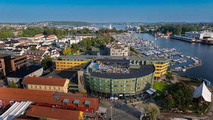 A green and yellow building with rooftop gatherings overlooks a marina filled with boats, surrounded by a coastal town with colorful houses and lush greenery under a clear blue sky.