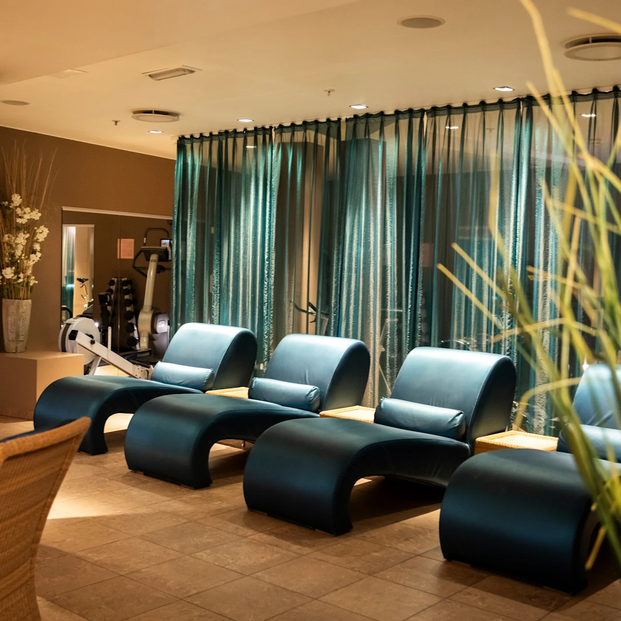 Five blue lounge chairs are neatly arranged in a room with warm lighting and tile flooring. In the background, there's a semi-transparent curtain, fitness equipment, and decorative plants.