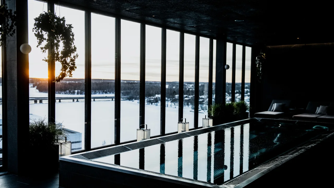 Inomhuspool med utsikt över solnedgången genom stora fönster som överblickar ett snötäckt landskap med en bro i fjärran.