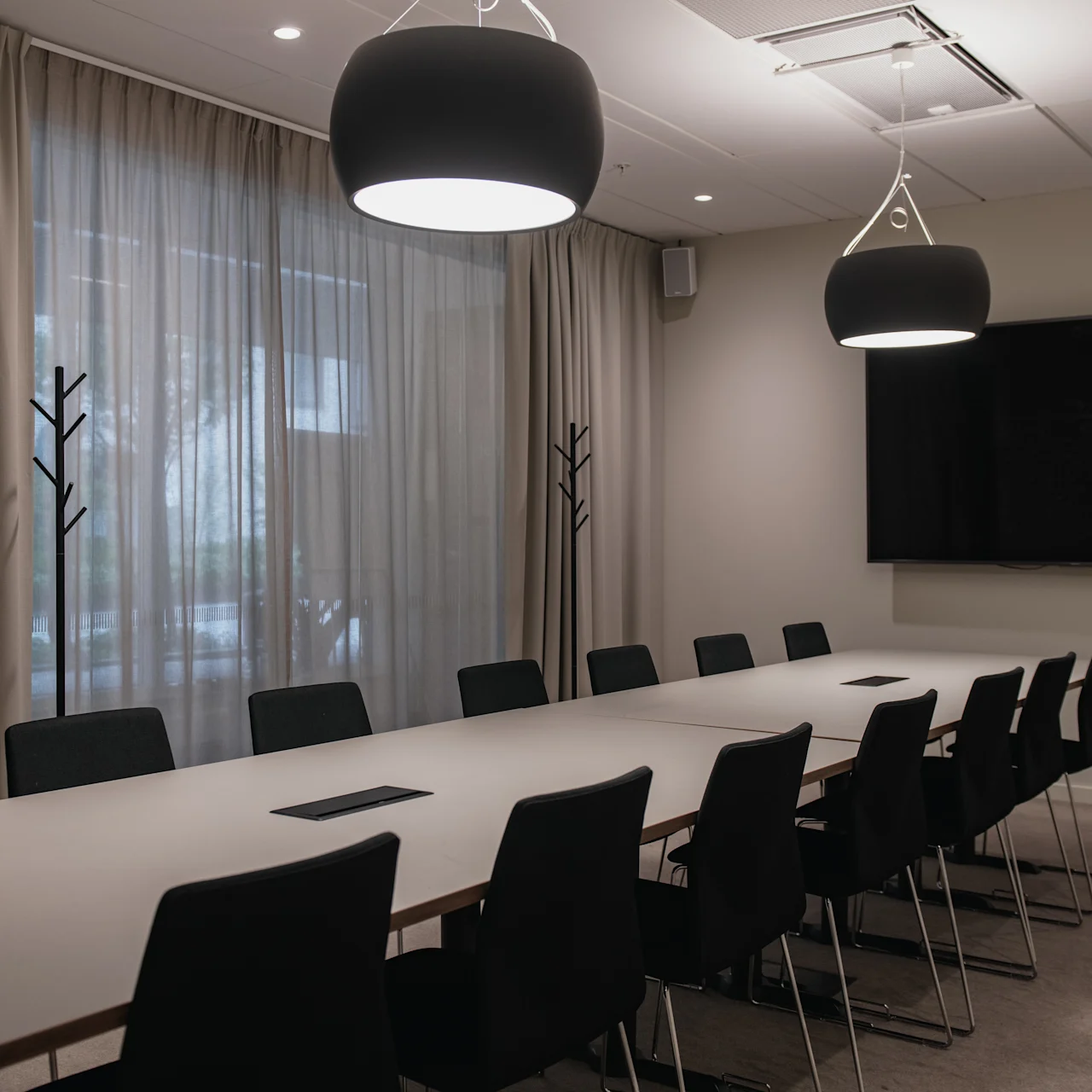 A long conference table with black chairs, situated in a modern meeting room. Overhead lights hang from the ceiling; a TV is mounted on the wall. Curtains cover the windows.