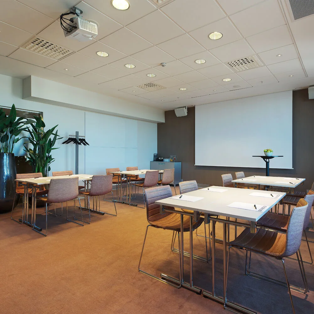 Conference room with tables arranged in a U-shape, chairs around them, and a projector on the ceiling. The room is well-lit and features a large screen and a plant.