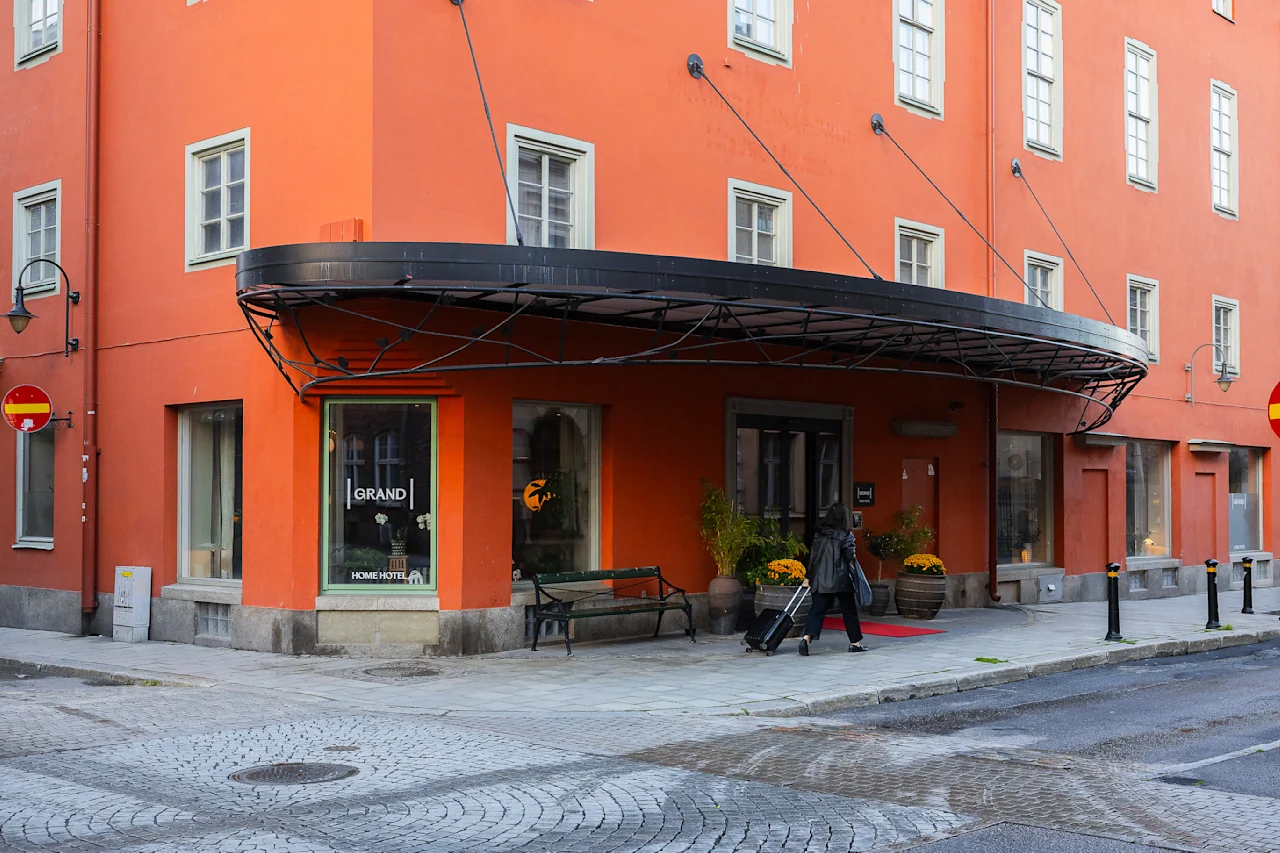 En person trækker en kuffert forbi indgangen til en orange bygning med et sort udhæng, mærket "GRAND HOME HOTEL." Gaden er brolagt med sten.
