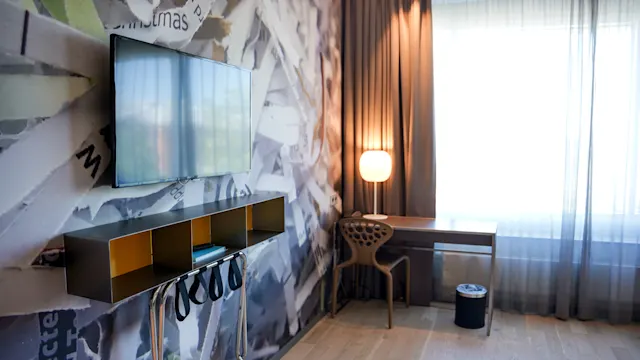 En tv är monterad på väggen, med en modern hylla under som visar föremål. Ett skrivbord och stol står bredvid en tänd lampa, intill ett fönster med skira gardiner.